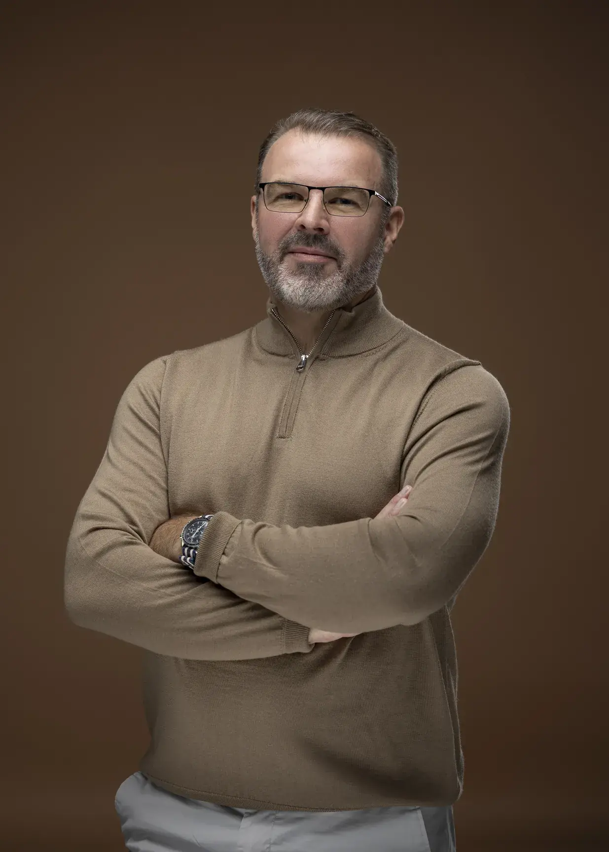 Rik Williams in a camel zip knit and glasses, arms crossed, brown backdrop. Corporate portrait photography by MRPS in Dubai, photographed by Nastaran.