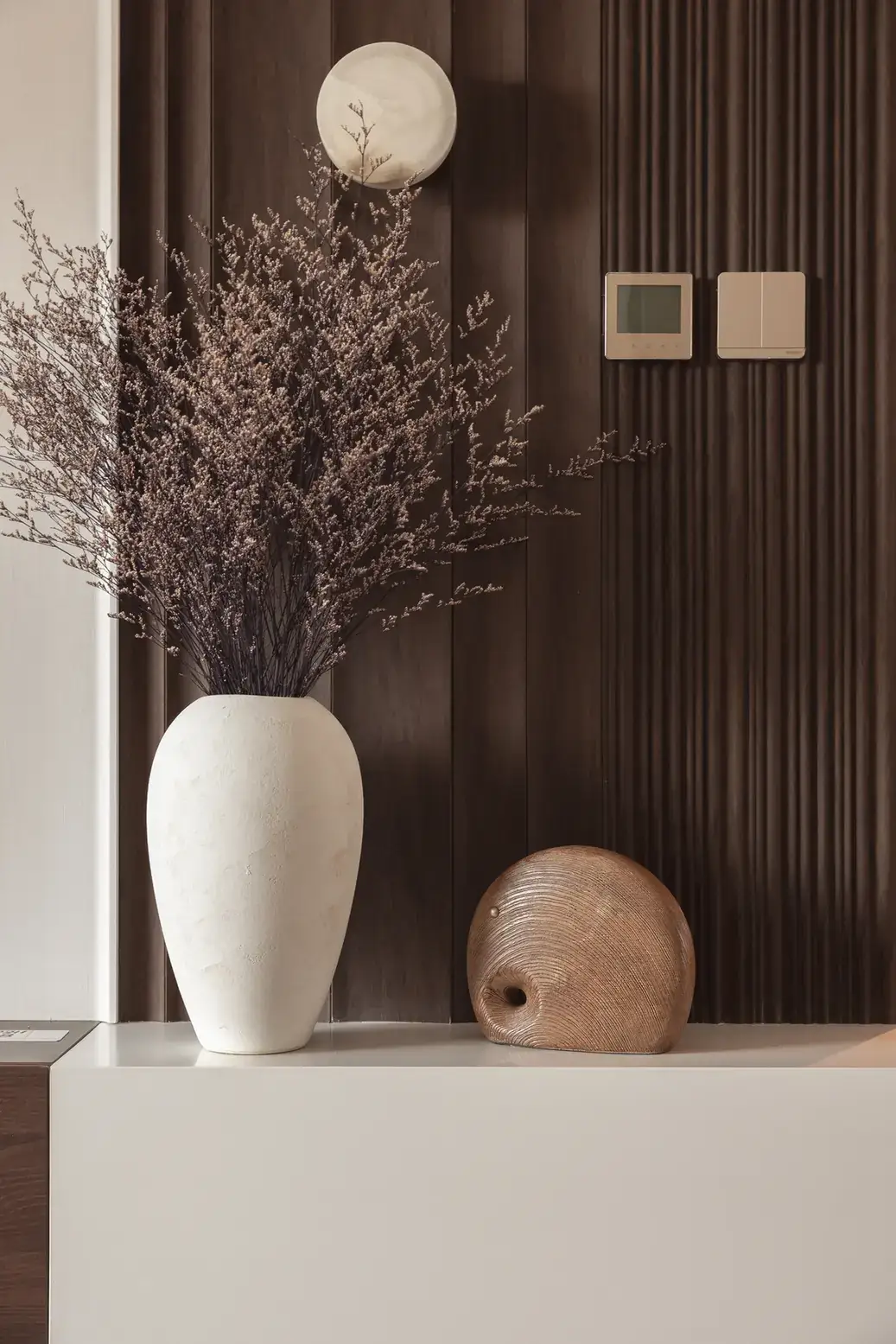Dried flowers in a white vase against a dark wooden slat wall panel. Interior design photography for Aziza Home by MRPS in Dubai.