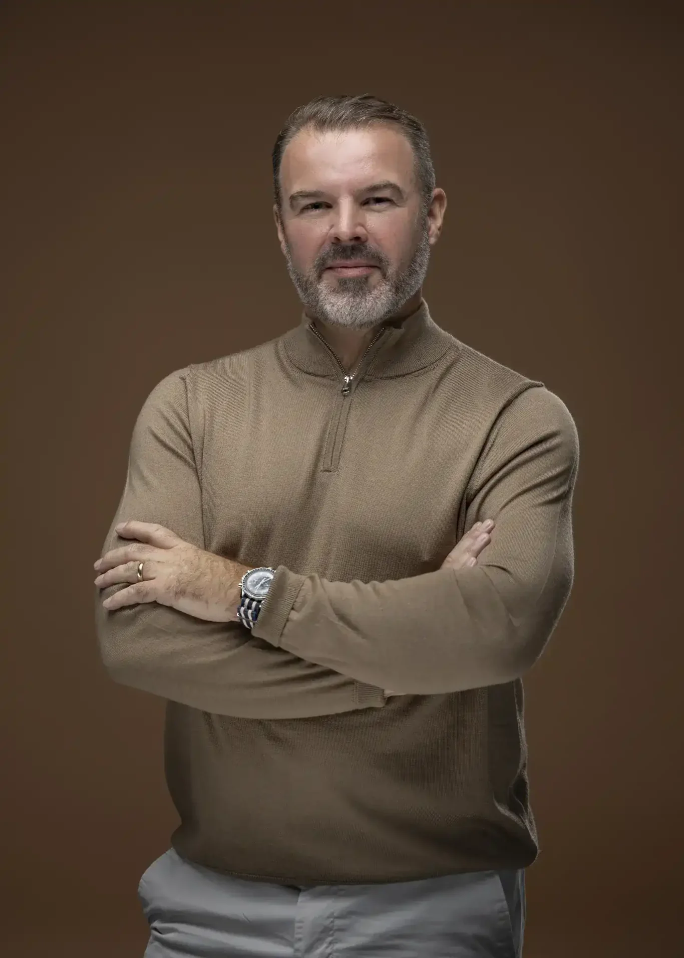 Rik Williams smiling in a camel zip knit with arms crossed, brown backdrop. Corporate portrait photography by MRPS in Dubai, photographed by Nastaran.