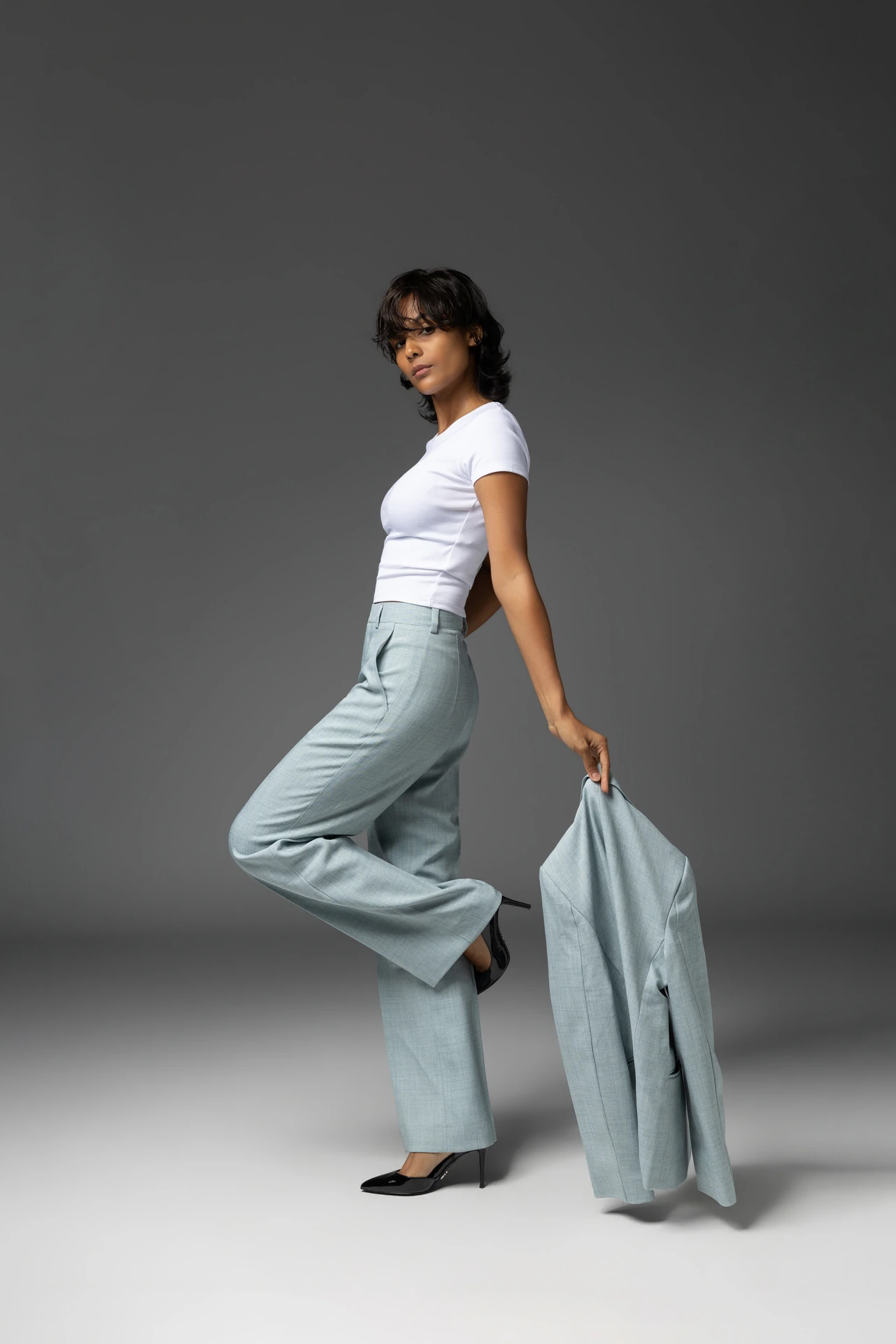 Mia in a white t-shirt and light blue tailored trousers, stepping forward while holding the matching blazer.