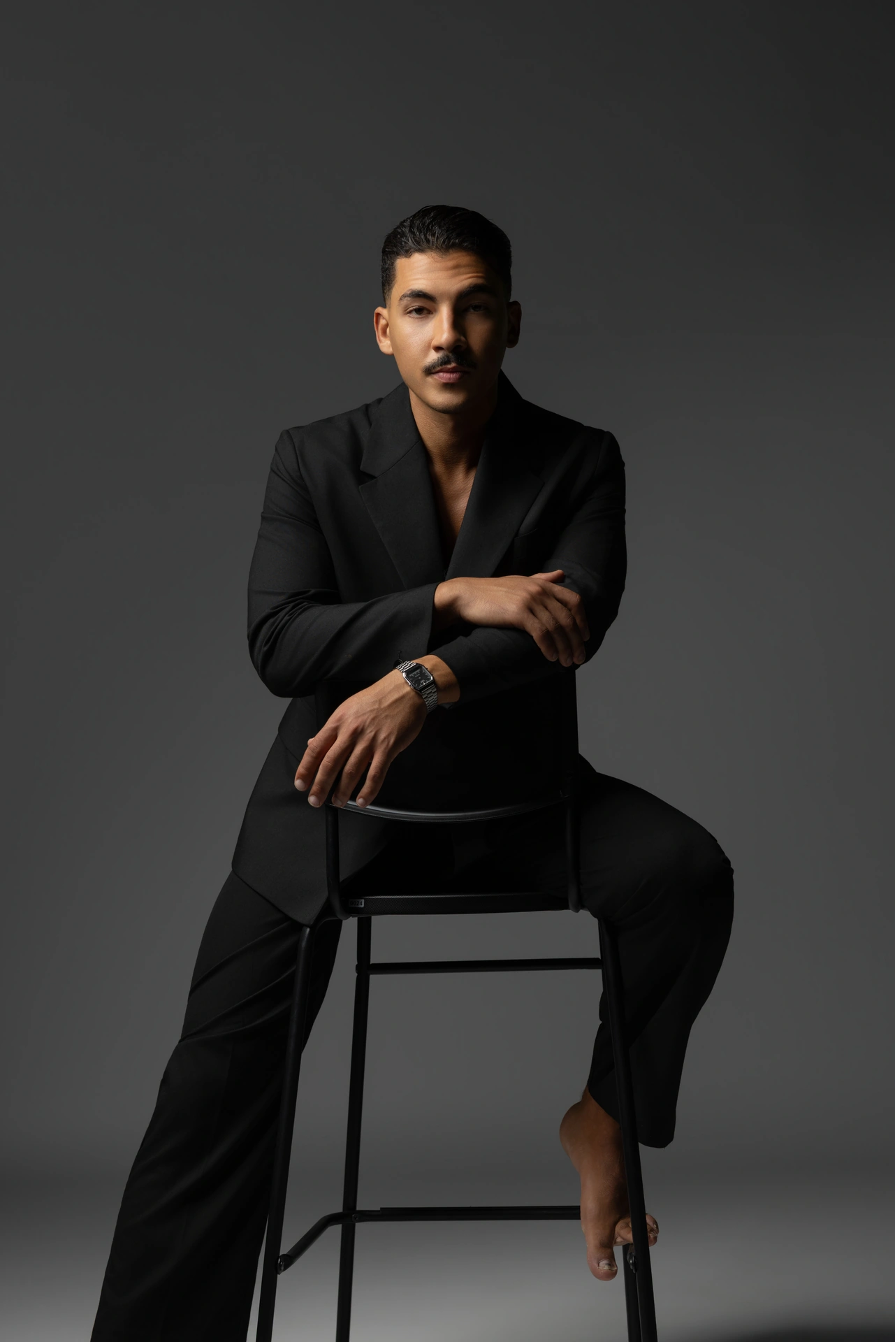 Seated on a stool in an open black blazer and black trousers. Male modeling portfolio photoshoot by MRPS in Dubai, photographed by Nastaran.