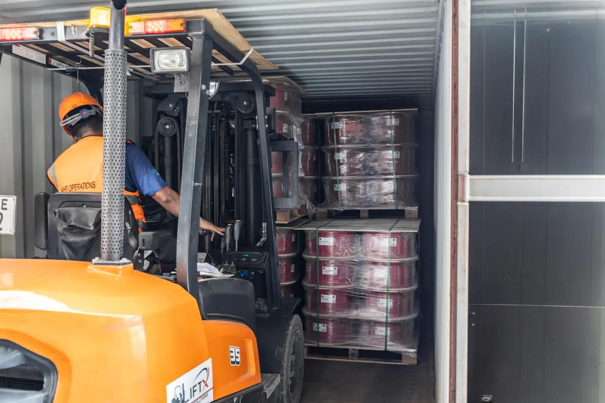 Forklift operator placing pallets of red cable reels inside a shipping container, seen from behind the forklift.