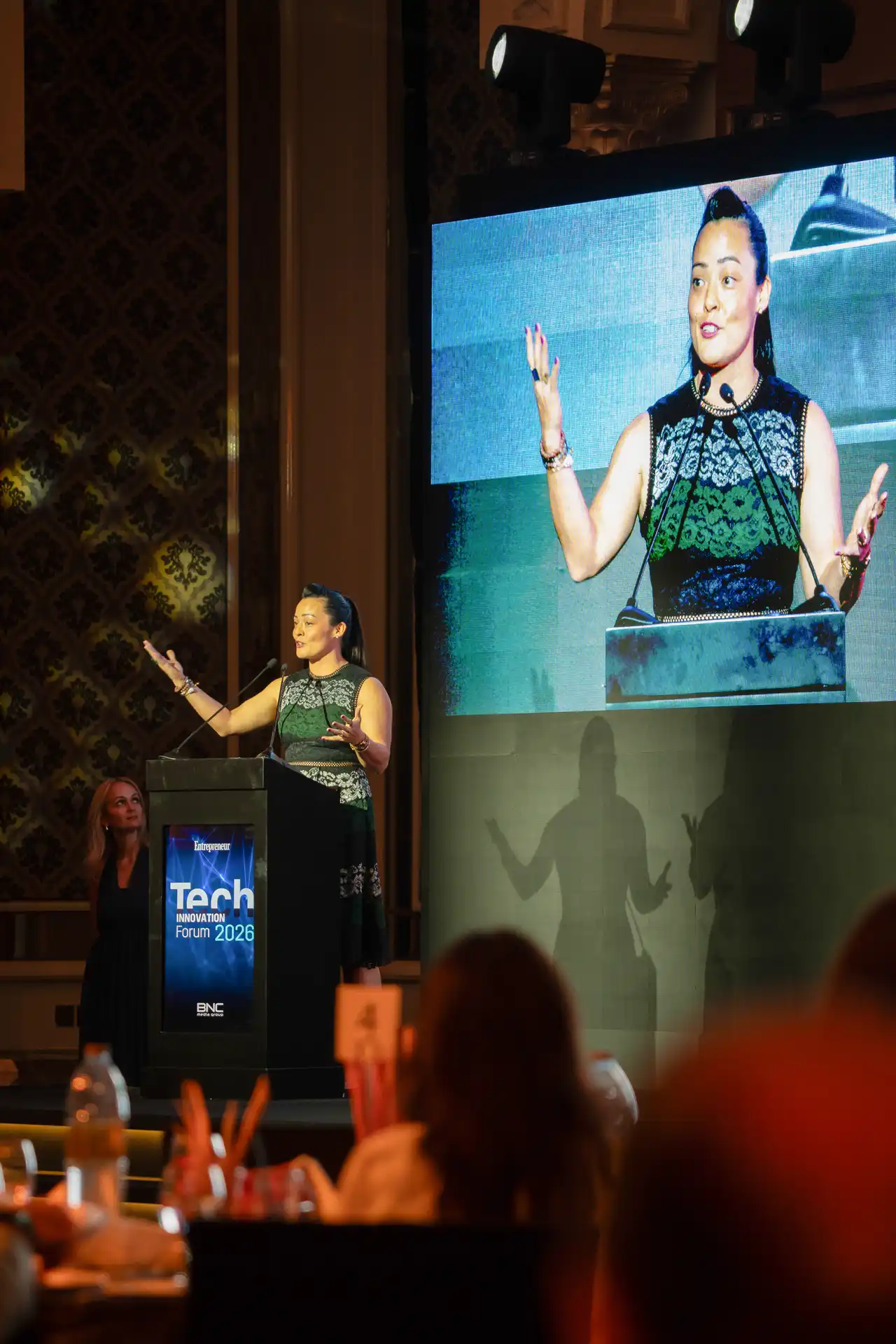 Speaker at the podium with her image on the big screen. Event photography by MRPS for FundedNext at the Tech Innovation Awards 2026, Al Habtoor Palace, Dubai.