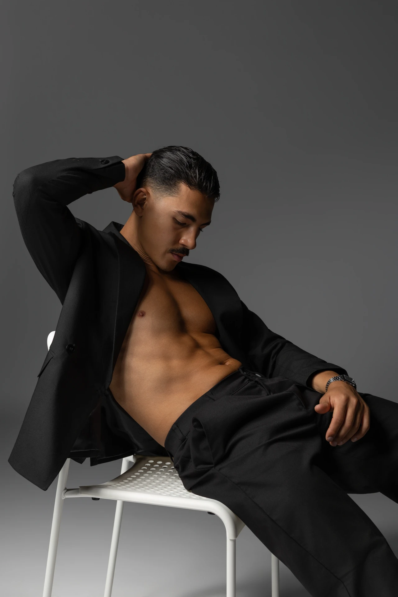 Relaxed seated pose on a white chair in an open black blazer, hand behind the head. Male modeling portfolio photoshoot by MRPS in Dubai, photographed by Nastaran.