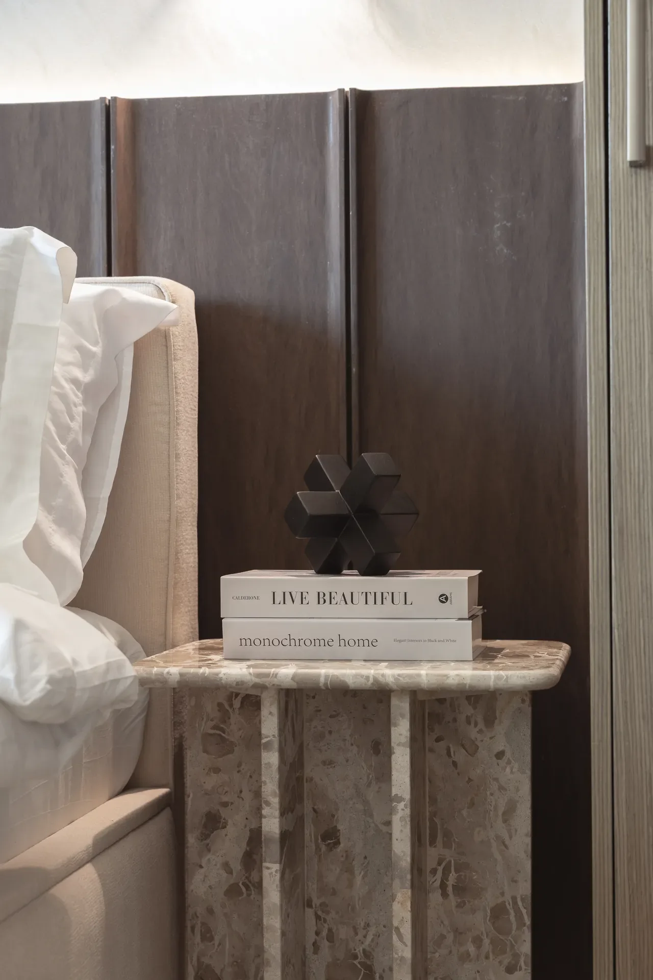 Travertine nightstand with books and a sculpture beside the bed. Interior design photography for Aziza Home by MRPS in Dubai.
