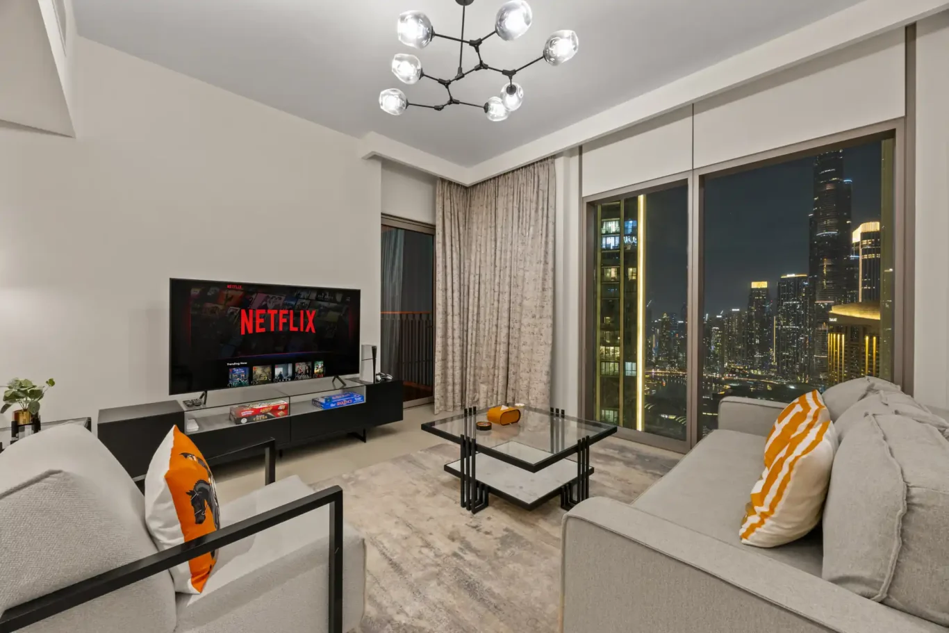 Grey sofa and orange cushions at twilight with the city lights outside. Day, twilight and night Airbnb photography by MRPS for Vista Haus Holiday Homes in Downtown Dubai.